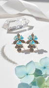 Pair of floral earrings with blue gemstones on a white surface with hydrangea flowers.