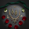 A premium multi-color American Diamond necklace and earring set with emerald and ruby tones, styled on a dark stone background with rose petals and green silk ribbon.