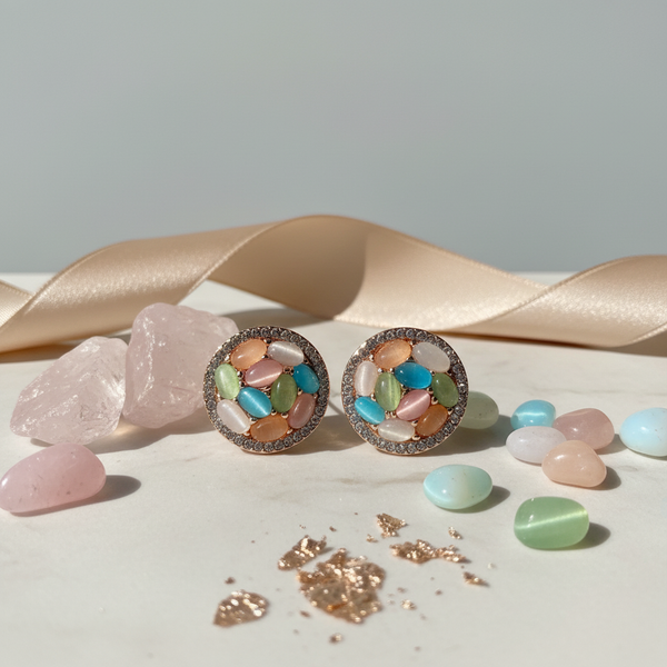 A pair of Diaura Jewels Korean style circular stud earrings featuring multi-color pastel stones on a white marble surface with rose quartz and silk ribbon.